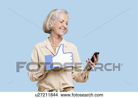 Picture - Senior woman with cell phone holding fake like button against blue background. Fotosearch
