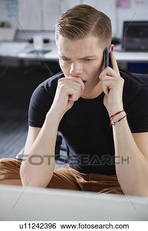 Serious young man looking at laptop while using cell phone in office View Large Photo Image Stock Photograph - Serious young man looking at laptop while using cell phone in office. Fotosearch