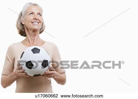 Stock Image - Smiling senior woman in casuals holding soccer ball against white background. Fotosearch