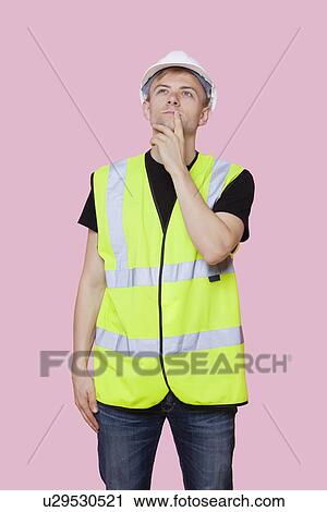 Stock Image - Thoughtful young construction worker looking away over pink background. Fotosearch