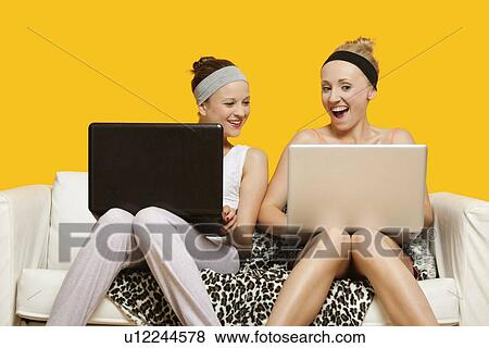 Two happy young women using laptop sitting on sofa against yellow background View Large Photo Image Stock Photo - Two happy young women using laptop sitting on sofa against yellow background. Fotosearch