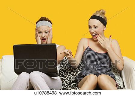 Stock Photo - Two shocked young women using laptop sitting on sofa against yellow background. Fotosearch