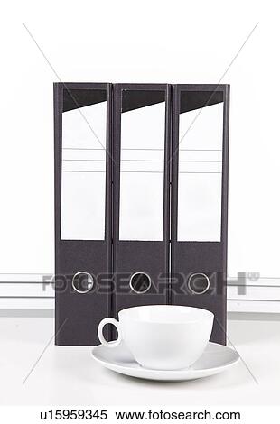Stock Photography - White coffee cup and saucer in front of folders at desk against white wall. Fotosearch