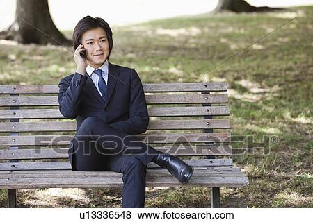 Young businessman using cell phone while sitting on bench at park View Large Photo Image Stock Photo - Young businessman using cell phone while sitting on bench at park. Fotosearch