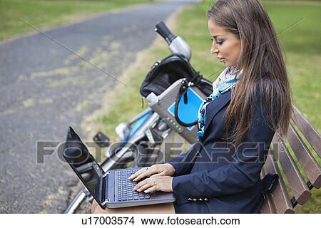 Picture - Young businesswoman using laptop while sitting on bench at park. Fotosearch