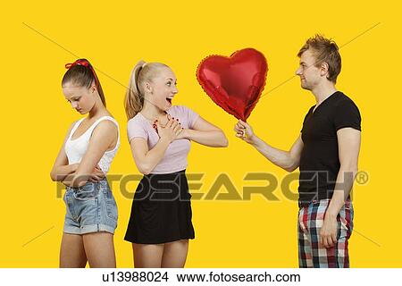 Young man gifting heart shaped balloon to surprised woman with friend feeling left out standing behind View Large Photo Image Picture - Young man gifting heart shaped balloon to surprised woman with friend feeling left out standing behind. Fotosearch