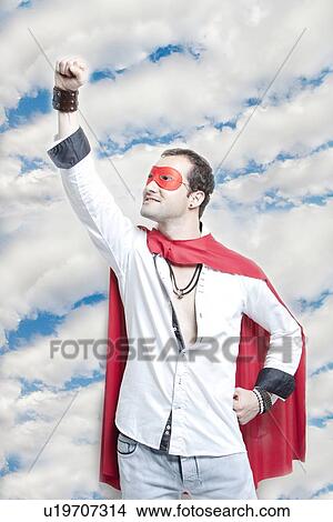 Picture - Young man in superhero costume with hand raised against cloudy sky. Fotosearch