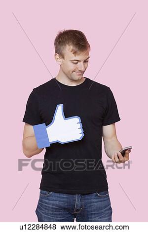 Young man with cell phone holding fake like button against pink background View Large Photo Image Stock Photo - Young man with cell phone holding fake like button against pink background. Fotosearch