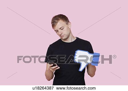 Young man with cell phone holding fake dislike button against pink background View Large Photo Image Stock Photo - Young man with cell phone holding fake dislike button against pink background. Fotosearch