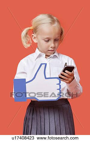 Stock Image - Young schoolgirl with cell phone holding fake like button against orange background. Fotosearch