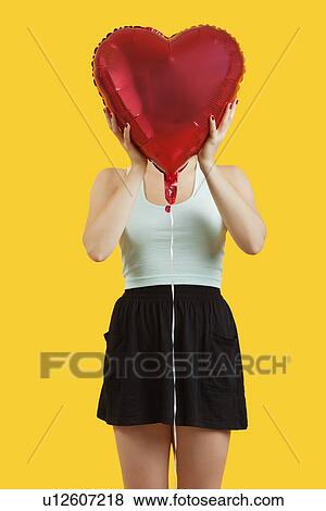 Young woman hiding behind heart shaped balloon over yellow background View Large Photo Image Stock Photo - Young woman hiding behind heart shaped balloon over yellow background. Fotosearch