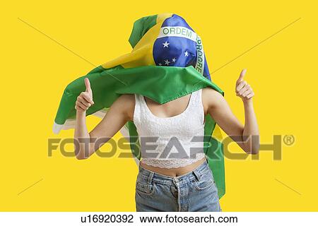 Stock Image - Young woman with Brazilian flag on face gesturing thumbs up over yellow background. Fotosearch