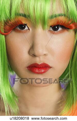 Stock Photo - Close-up portrait of young woman wearing green wig and orange eyelashes over gray background. Fotosearch