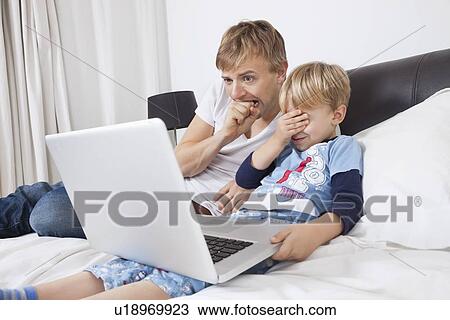 Stock Image - Father and son watching scary movie on laptop in bed. Fotosearch