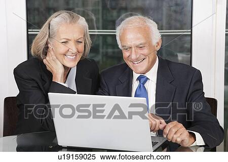 Stock Photography - Happy senior business couple looking at laptop white sitting at table. Fotosearch