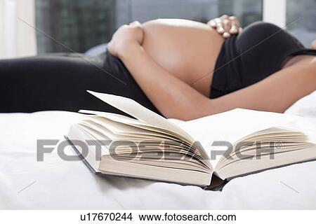 Mid section of pregnant woman lying in bed with open book in foreground View Large Photo Image Picture - Mid section of pregnant woman lying in bed with open book in foreground. Fotosearch