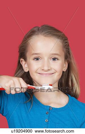 Stock Image - Portrait of girl holding toothbrush against red background. Fotosearch