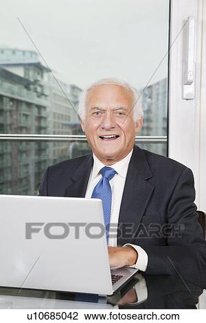 Stock Image - Portrait of happy elderly businessman with laptop sitting at table. Fotosearch