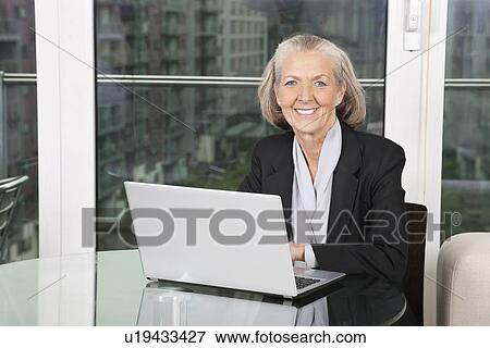Portrait of happy senior businesswoman with laptop sitting at table View Large Photo Image Stock Photo - Portrait of happy senior businesswoman with laptop sitting at table. Fotosearch