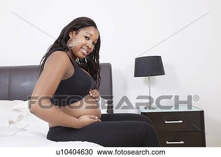 Stock Image - Portrait of happy young pregnant woman touching her belly in bed. Fotosearch