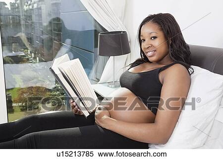 Stock Photography - Portrait of happy young pregnant woman reading book on sofa. Fotosearch