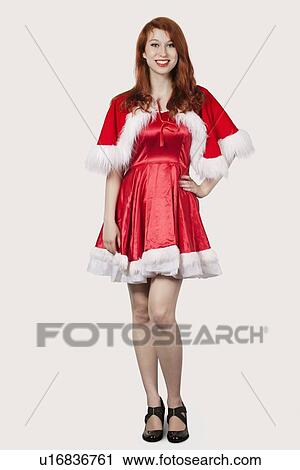 Stock Image - Portrait of happy young woman in Santa costume standing against gray background. Fotosearch