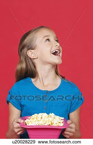Stock Photo - Portrait of surprised young girl holding popcorn container against red background. Fotosearch