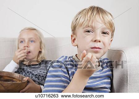 Stock Image - Portrait of young boy with sister watching TV and eating popcorn. Fotosearch