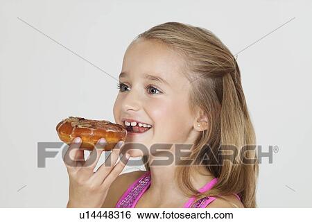 Stock Photograph - Portrait of young girl eating donut against gray background. Fotosearch