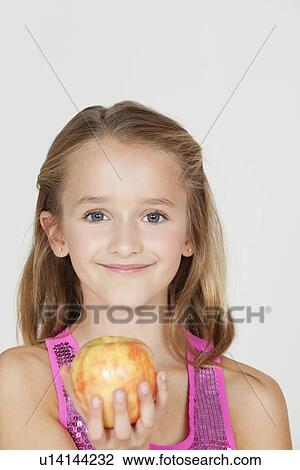 Stock Image - Portrait of young girl holding an apple against gray background. Fotosearch
