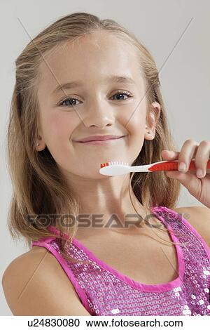 Stock Image - Portrait of young girl holding toothbrush against gray background. Fotosearch