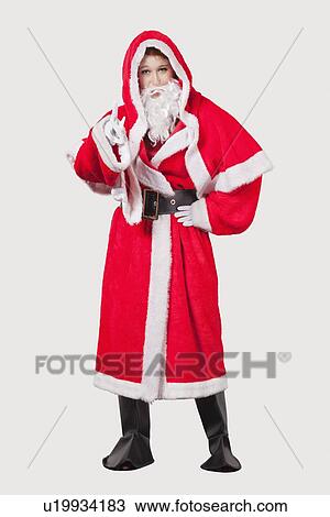 Stock Image - Portrait of young woman in Santa costume gesturing against gray background. Fotosearch