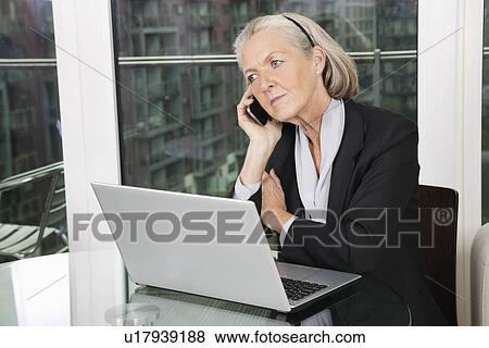 Senior businesswoman with laptop using cell phone while sitting at table View Large Photo Image Stock Photo - Senior businesswoman with laptop using cell phone while sitting at table. Fotosearch