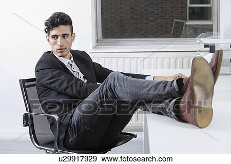 Stock Photo - Thoughtful young businessman sitting with legs raised at desk in office. Fotosearch