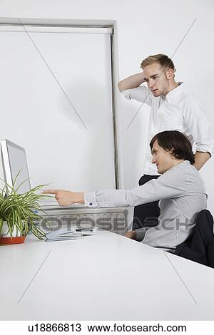 Stock Image - Young businessman showing computer screen to confused coworker at desk. Fotosearch