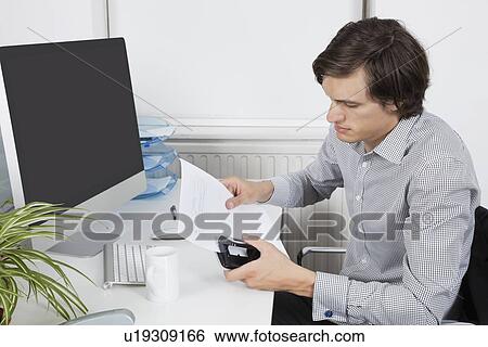 Stock Photograph - Young businessman using hole puncher at desk. Fotosearch