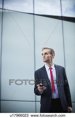Young businessman with cell phone standing against glass building View Large Photo Image Stock Image - Young businessman with cell phone standing against glass building. Fotosearch