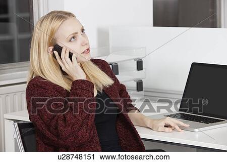 Stock Image - Young businesswoman using cell phone while sitting at desk in office. Fotosearch