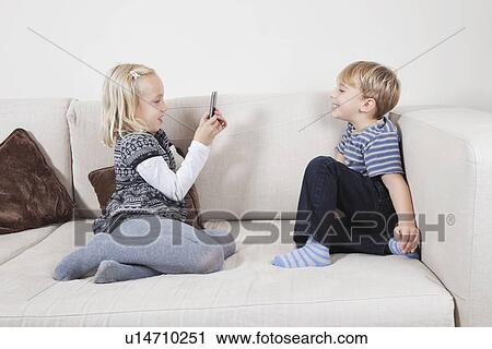 Young girl photographing brother through cell phone on sofa View Large Photo Image Stock Image - Young girl photographing brother through cell phone on sofa. Fotosearch