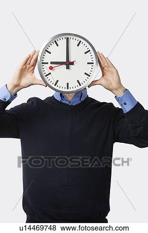 Stock Photo - Young man covering his face with clock standing against white background. Fotosearch
