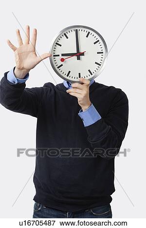 Stock Photo - Young man holding clock over his face gesturing against white background. Fotosearch