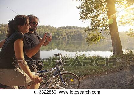 Stock Image - Couple riding bicycles by river bank. Fotosearch