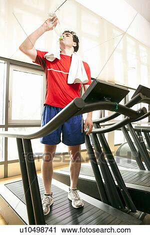 Stock Image - Man drinking water on treadmill at gym. Fotosearch