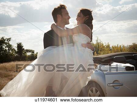 Stock Image - Newlywed groom carrying bride outdoors. Fotosearch