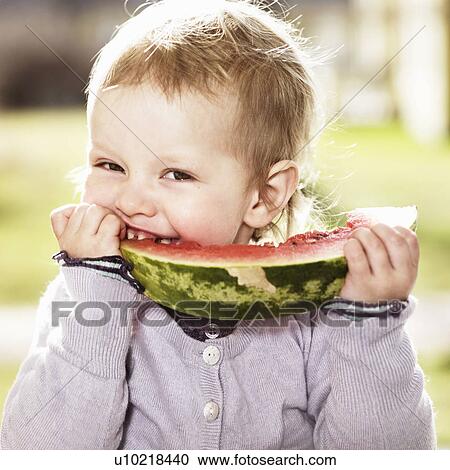 Stock Image - Toddler girl eating watermelon outdoors. Fotosearch