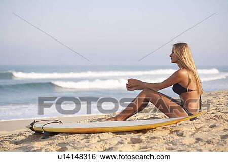 Stock Photograph - Woman sitting with surfboard on beach. Fotosearch