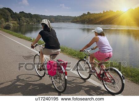 Stock Photography - Women riding bicycles by river bank. Fotosearch