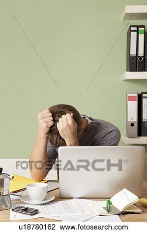 Stock Image - Anxious man tearing at hair at desk. Fotosearch