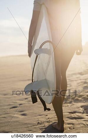 Stock Image - Female surfer walking on beach at sunset. Fotosearch