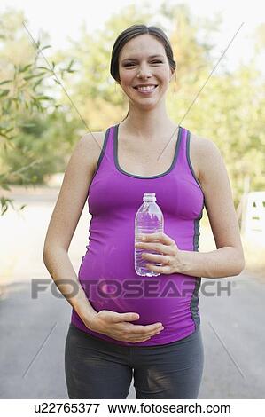 Stock Photo - Front view portrait of pregnant mid adult woman in sport clothing holding water bottle. Fotosearch